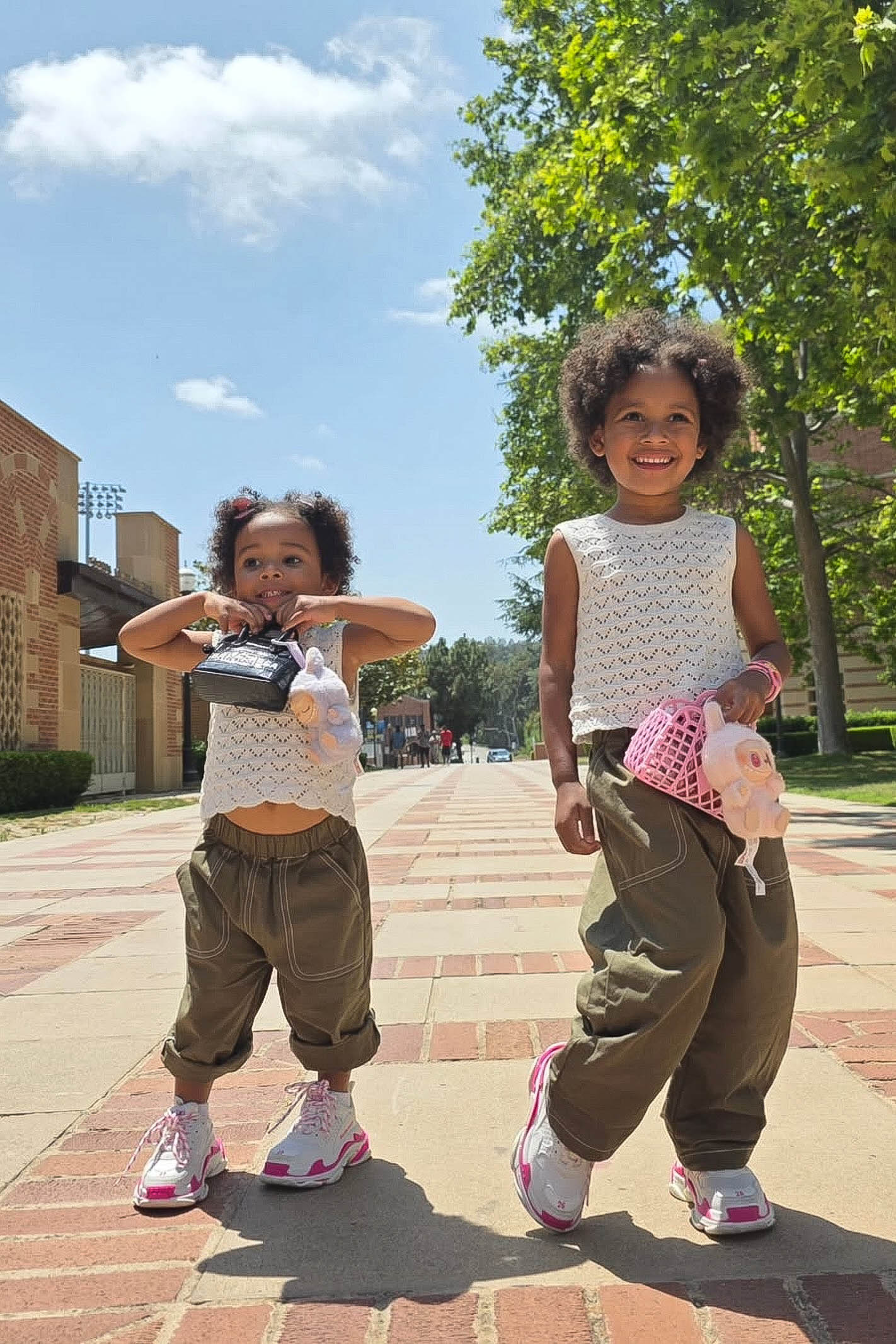 Two young girls standing on a brick sidewalk with trees and buildings in the background.