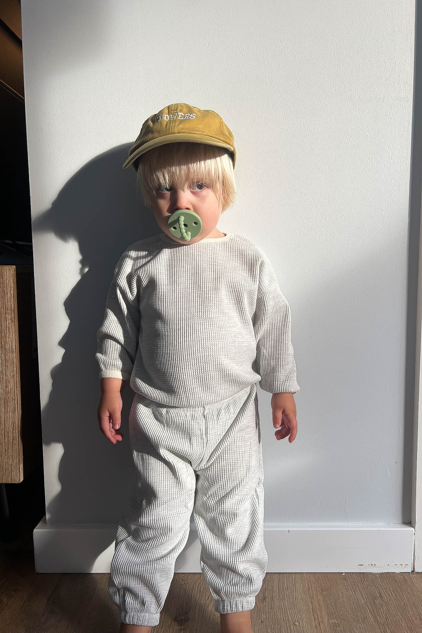 Child wearing a cap and light-colored outfit against a plain wall.