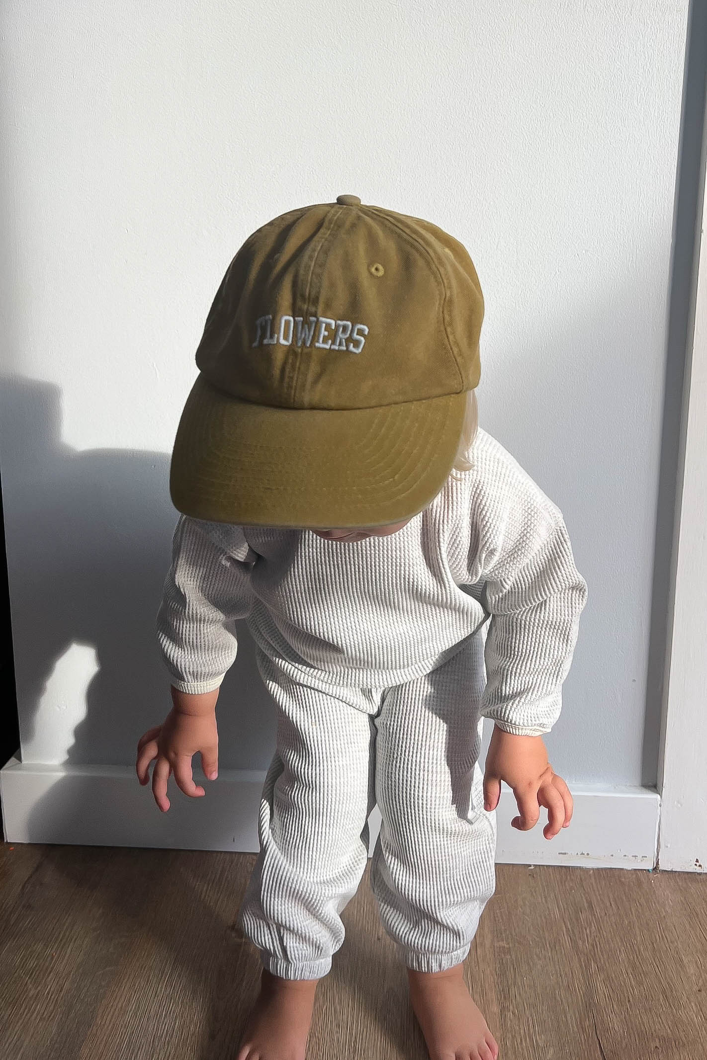 Child wearing a green cap with &#39;FLOWERS&#39; text against a white wall.