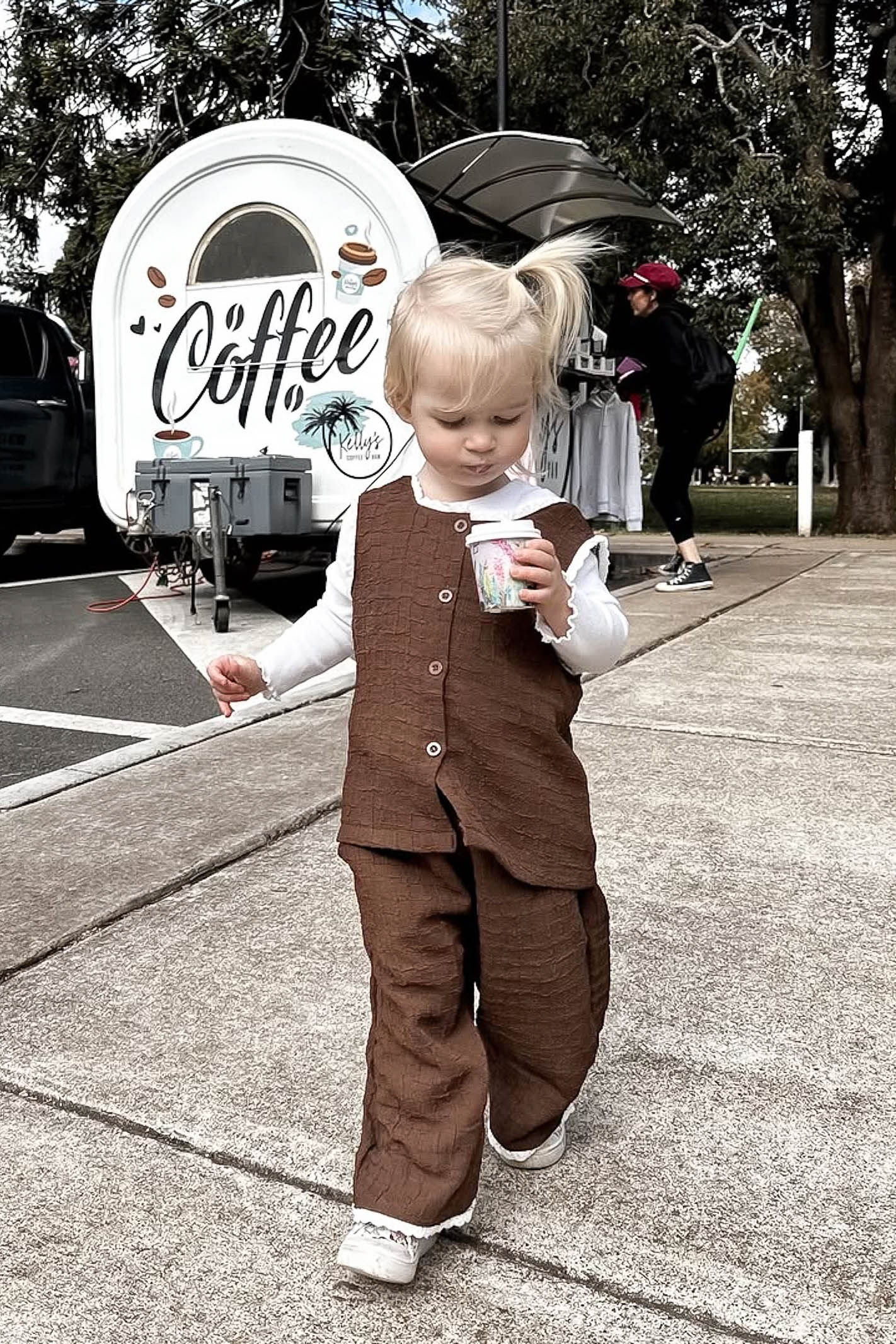 Child in a brown outfit walking on a sidewalk with a coffee truck in the background.