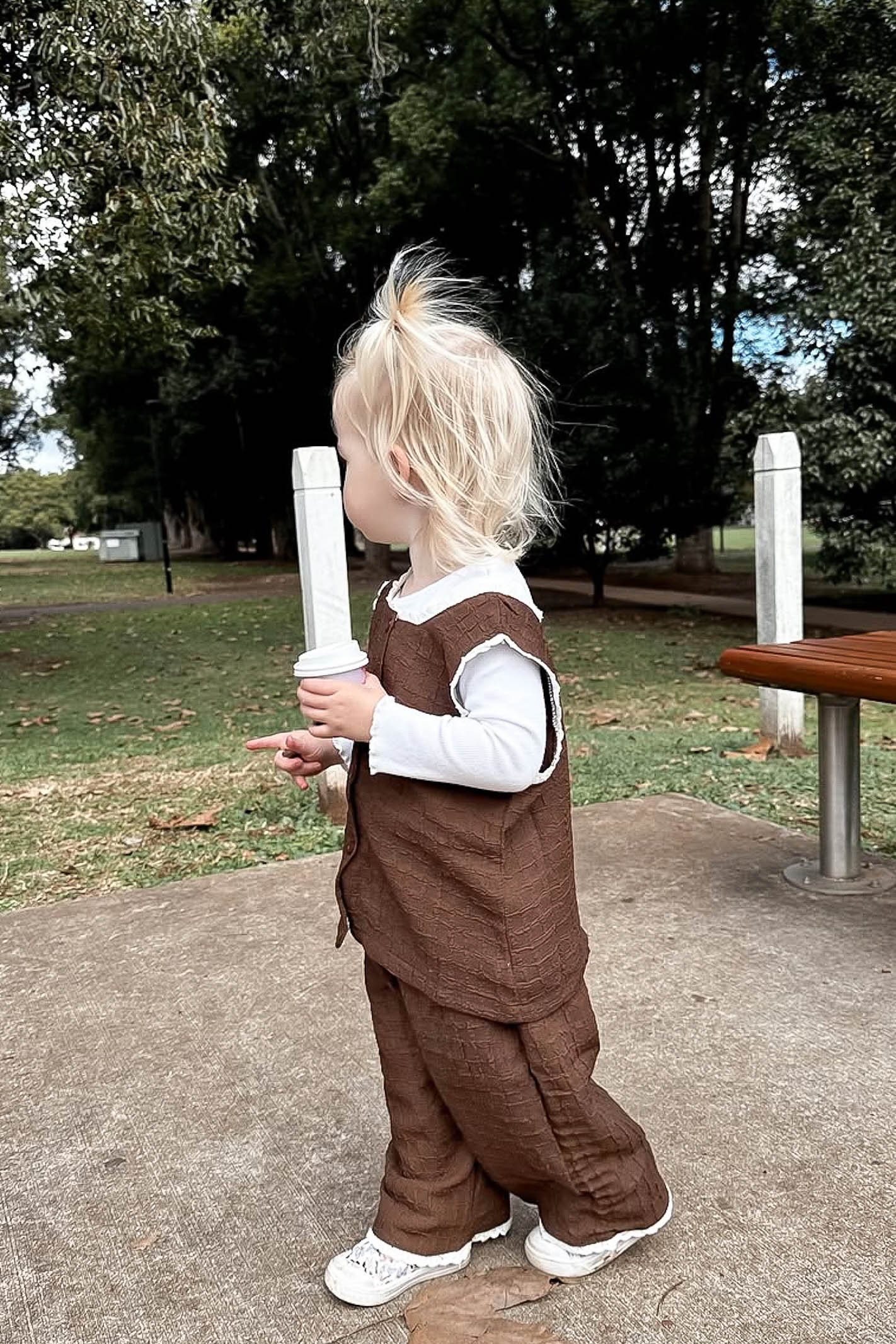 Child in a brown outfit holding a white object outdoors with trees in the background