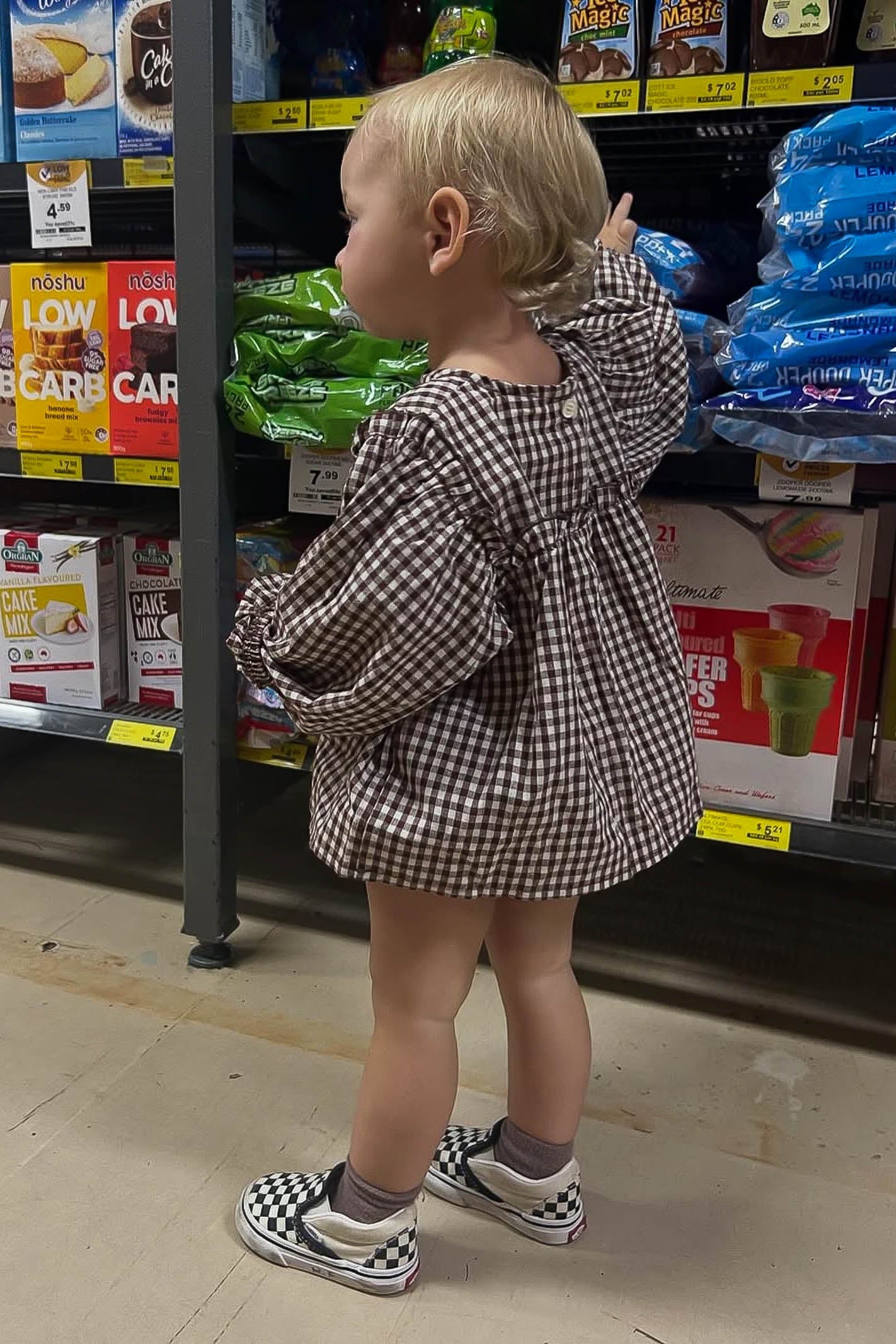 Child in a gingham outfit standing in front of a grocery store shelf.
